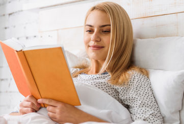 Happy young woman is reading book in bed with pleasure. She is sitting and smiling