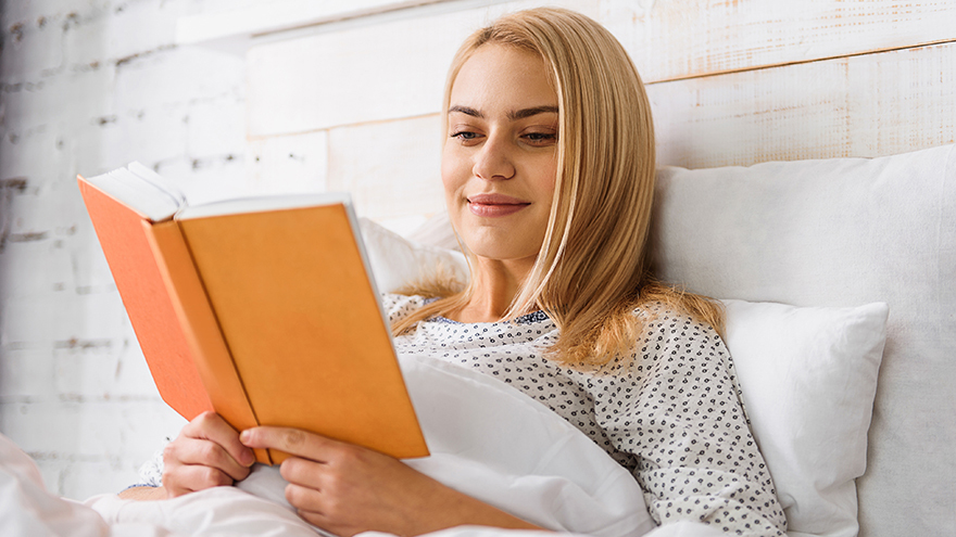 Happy young woman is reading book in bed with pleasure. She is sitting and smiling