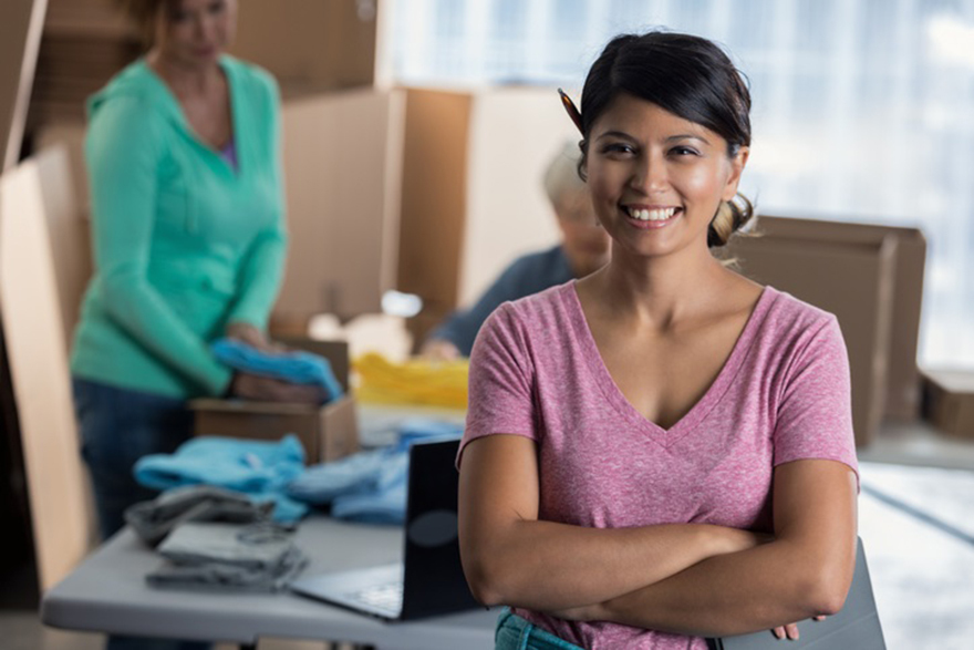 A mid adult small business entrepreneur confidently stands in distribution warehouse. Her business partners are working in the background. She is standing with her arms crossed.