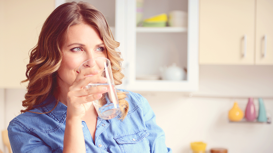 Blonde woman in blue blouse drinking glass of water in kitchen