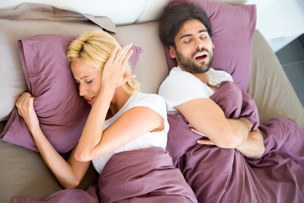 Young couple lying in bed. Young woman is very irritated by her boyfriends snoring and covering her ears with her hands. High angle view.