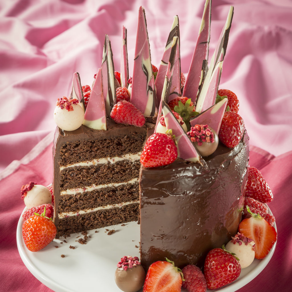Ultimate Chocolate Cake decorated with strawberries, chocolates and multi coloured chocolate shards,a big slice cut showing 3 layers of cream