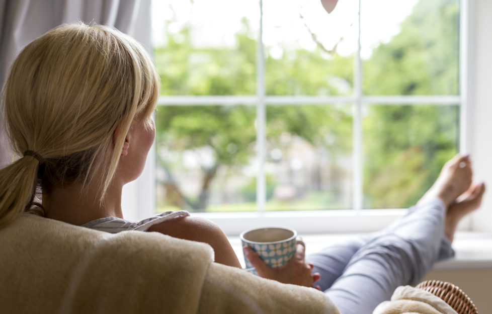 A young woman sits with a hot drink and her feet up, watching out of the window from the comfort of her own home. She is taking a break from her chores and busy day to day routine to gaze into her garden and wonder.