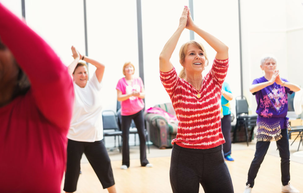 Women clap their hands over their heads as they learn new dance moves in thier local senior center. They are smiling and enjoying themselves.