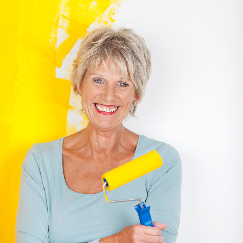 Smiling mature woman with paint roller, wall behind her is half painted yellow