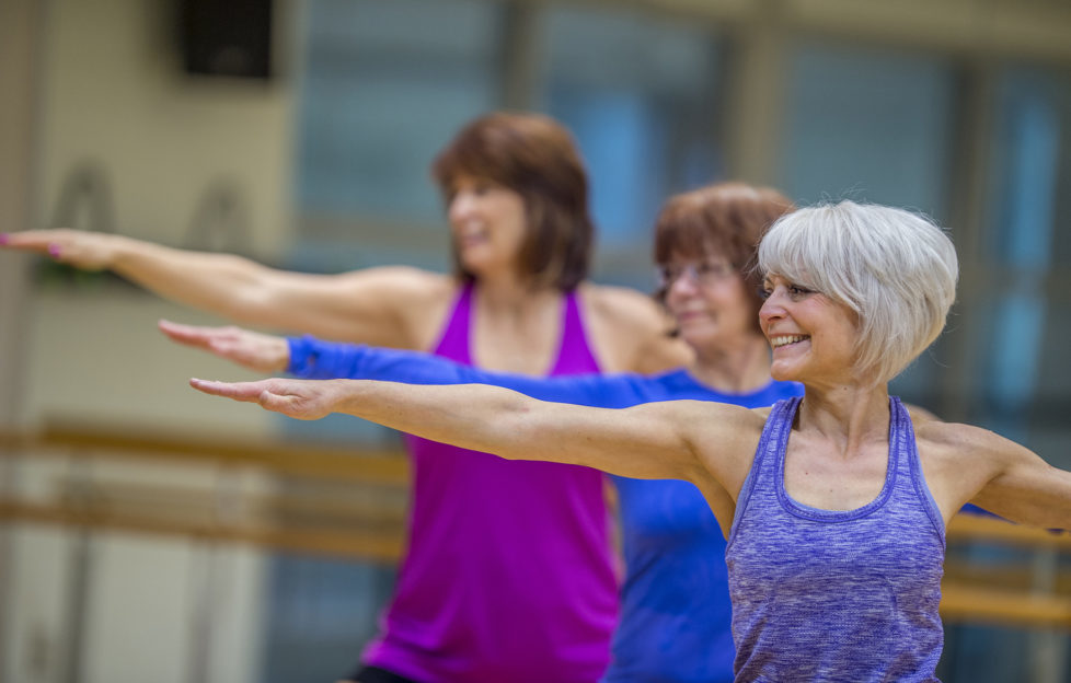 Three Caucasian senior women are indoors in a fitness center. They are wearing casual exercise clothing. They are standing and stretching their arms out while doing yoga. One woman is smiling in the foreground.