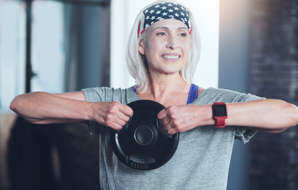 Getting stronger. Happy woman smiling while lifting a weight disk while doing swing exercise while having a training session indoors.