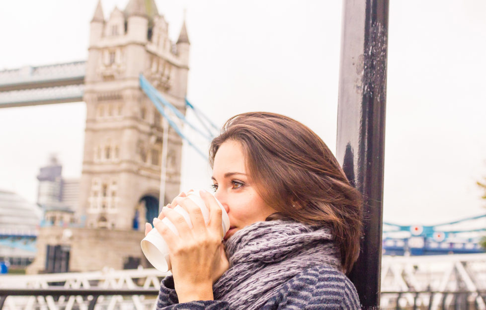 Young woman in a chilly autumn with a soft scarf, warms up drinking from coffee cup.Tower Bridge in the background