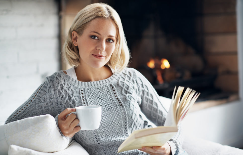 A young woman enjoying her novel with a hot drink while sitting inside near a fireplace Pic: Istockphoto