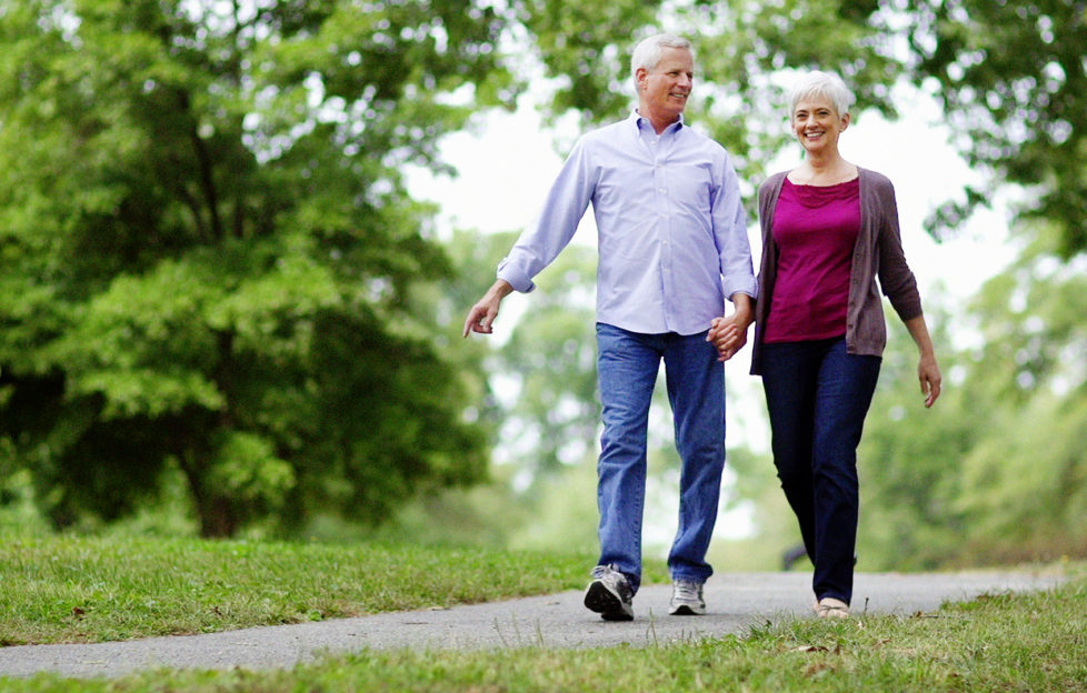 Senior couple walking in park on path with trees in the background.