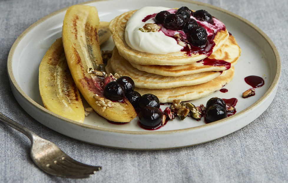 Small stack of pancakes on a plate with butter-fried bananas, lightly cooked blueberries and Greek yogurt