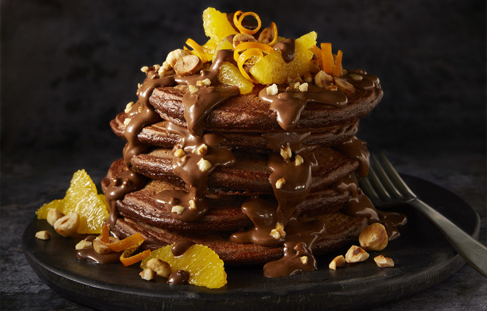 Stack of chocolate pancakes, drizzled with chocolate sauce, hazelnuts and garnished with orange segments