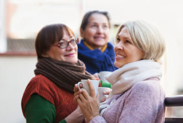 Senior female in warm clothes having cup of hot tea on terrace. Focus on blonde woman
