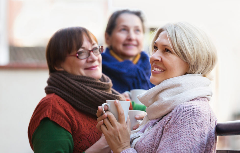 Senior female in warm clothes having cup of hot tea on terrace. Focus on blonde woman