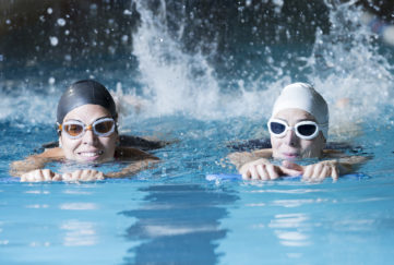couple of female swimmers swimming with a swim board doing leg exercises in an indoor swimming pool