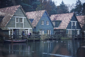 Three Scandinavian wooden houses, stained in green and blue with wide reddish tiled roofs, set beside a lake, family with children on the veranda wearing warm clothes