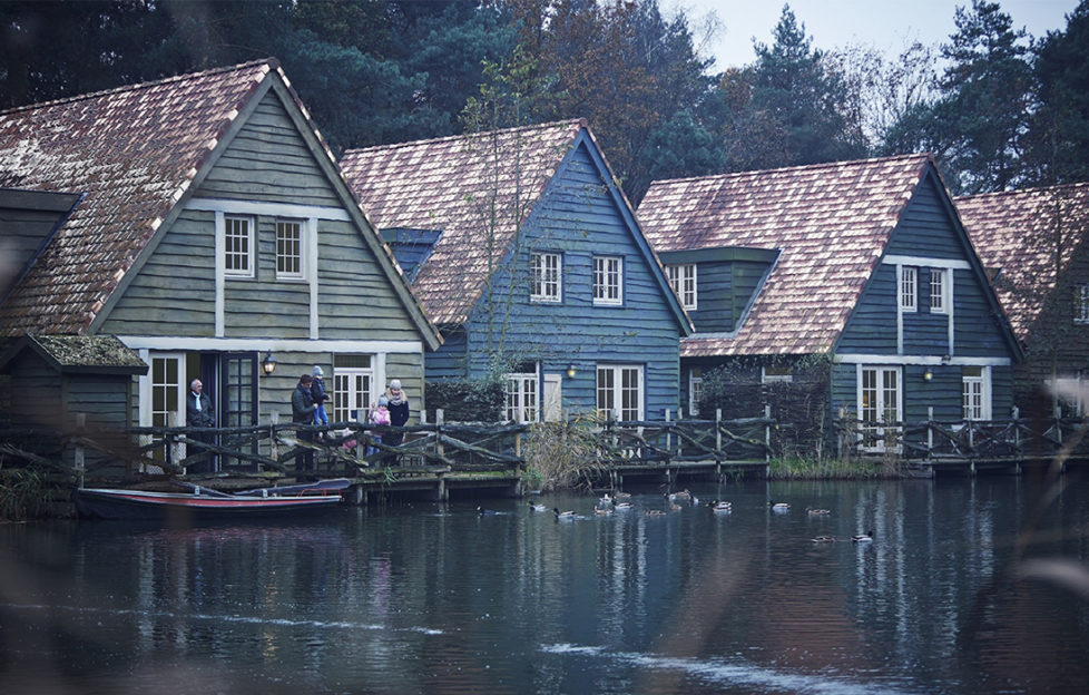 Three Scandinavian wooden houses, stained in green and blue with wide reddish tiled roofs, set beside a lake, family with children on the veranda wearing warm clothes