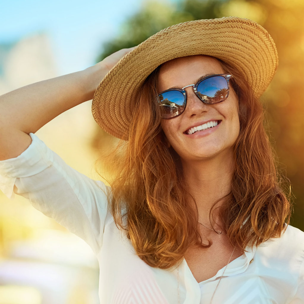 Auburn-haired woman in sunglasses, light shirt and floppy hat, enjoying a summer’s day outdoors