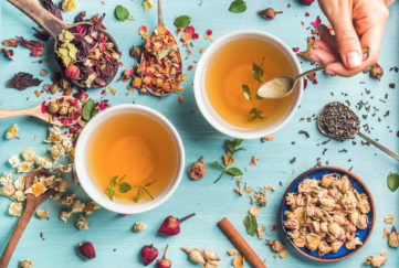Two cups of healthy herbal tea with mint, cinnamon, dried rose and camomile flowers in spoons and man's hand holding spoon of honey, blue background, top view