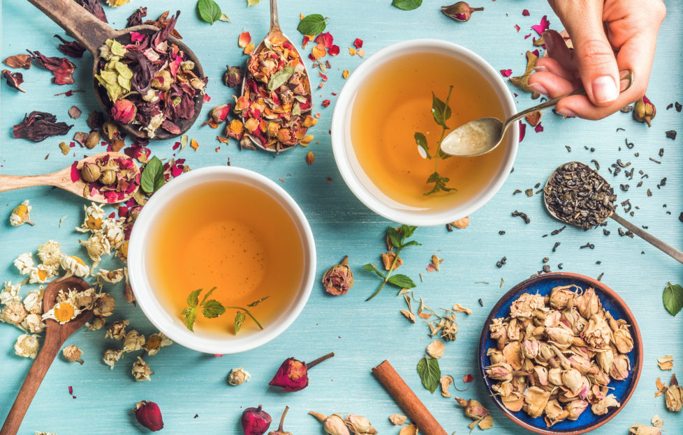 Two cups of healthy herbal tea with mint, cinnamon, dried rose and camomile flowers in spoons and man's hand holding spoon of honey, blue background, top view