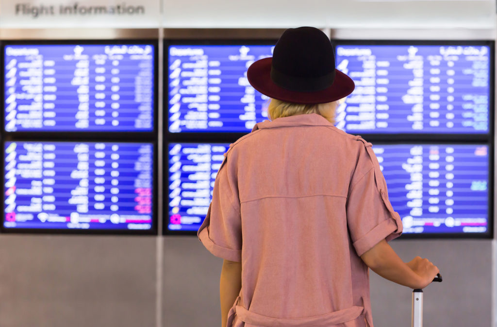 Young pretty woman in international airport looking at the flight information board with carry on luggage