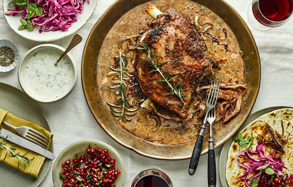 Roasted lamb in rich sauce with rosemary sprigs. Also glasses of red wine, pomegranate seeds, red cabbage and rosemary yogurt