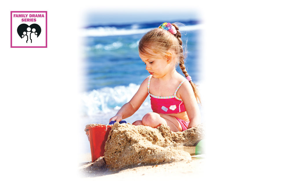 Little girl sitting with bucket and sandcastle, sea behind, long blonde plait and red bikini