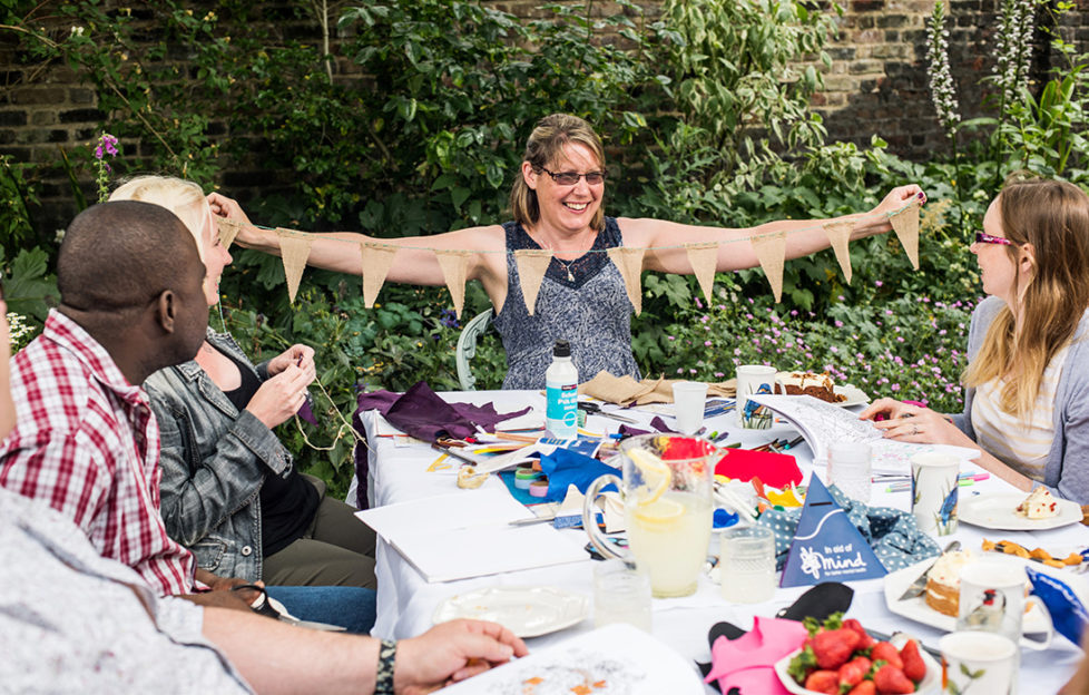 People sitting around a table covered in craft materials at a Crafternoon event. One woman is proudly holding up a string of home made bunting.