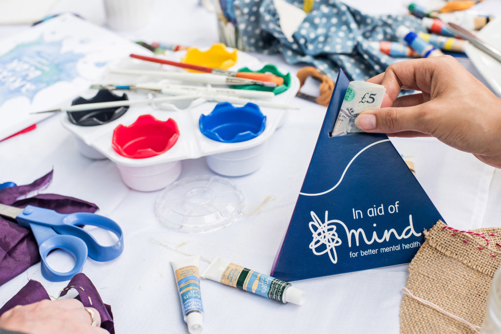 Table strewn with craft materials. A hand is posting £5 into a pyramid shaped Mind donations box.