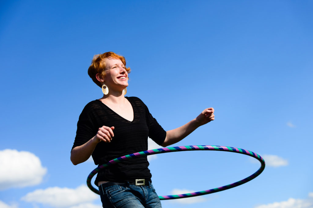 A red headed female outside playing with a hula hoop. Sky, clouds and trees in the background.