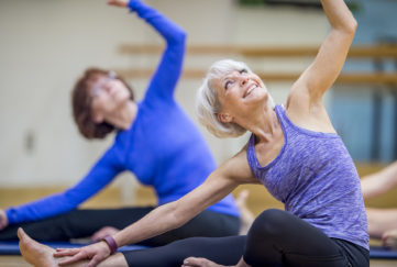 Two Caucasian women are indoors in a health center. They are wearing casual athletic clothing. They are sitting on the floor and reaching their arms into the air.