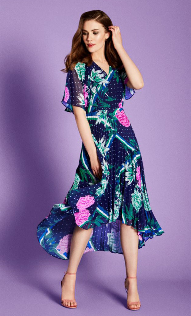 Brunette model in v neck floaty cap sleeve dress, navy with bright pink flowers and teal leaves, skirt of narrow pleats, hemline higher at the front