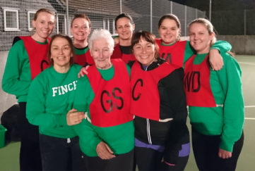 80-year-old netballer Anne Wilby with fellow members of her netball team, in netball vests on a floodlit outdoor court