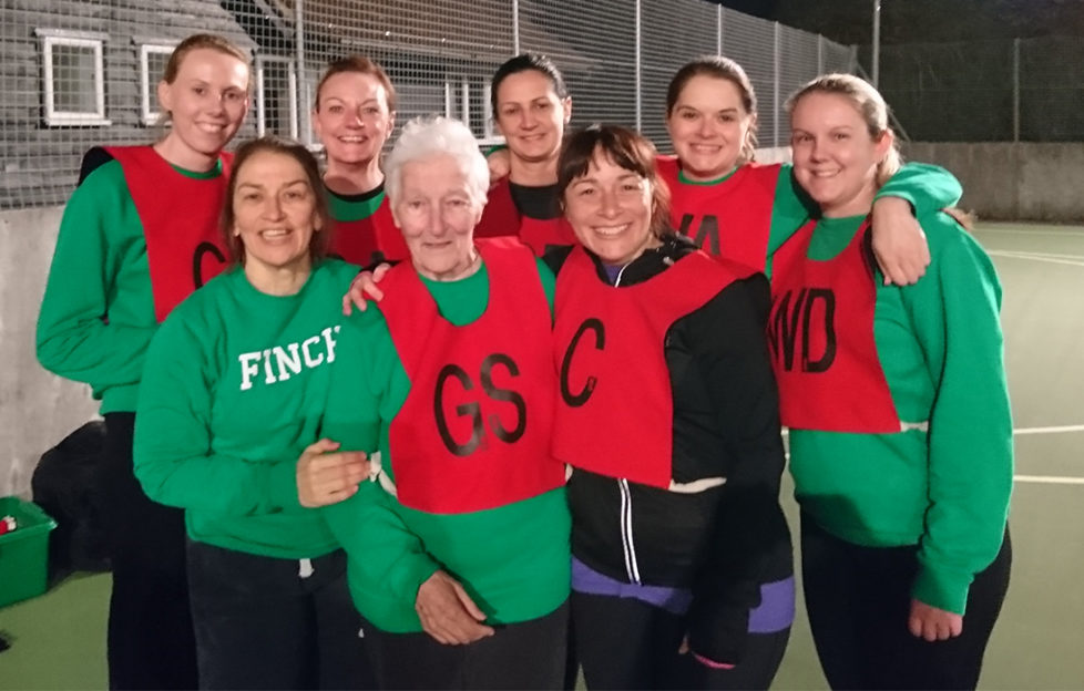 80-year-old netballer Anne Wilby with fellow members of her netball team, in netball vests on a floodlit outdoor court