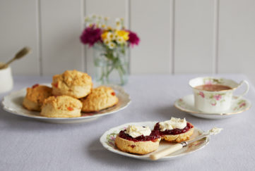 Plate of cherry scones, and another plate with a scone cut in half spread with jam and cream, also vase of flowers and teacup