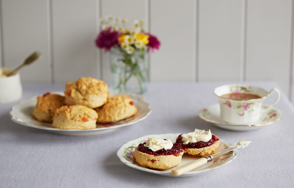 Plate of cherry scones, and another plate with a scone cut in half spread with jam and cream, also vase of flowers and teacup