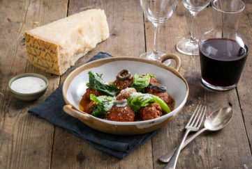 Bowl of four meatballs in tomato sauce, with finely grated grana padano cheese and wilted leaves, and a glass of red wine