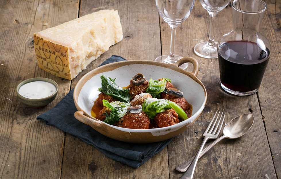 Bowl of four meatballs in tomato sauce, with finely grated grana padano cheese and wilted leaves, and a glass of red wine