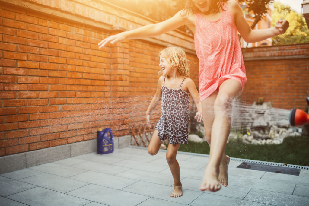 Two cute girls having fun in their backyard with garden hose. Jumping and running with arms raised.