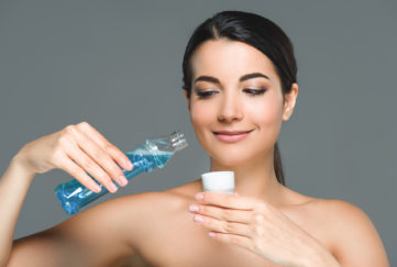 portrait of smiling woman with bare shoulders holding mouthwash isolated on grey