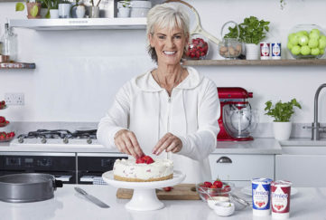 Judy Murray in kitchen, smiling, with strawberry cheesecake on worktop and plants, strawberries and jar of tennis balls on shelves behind