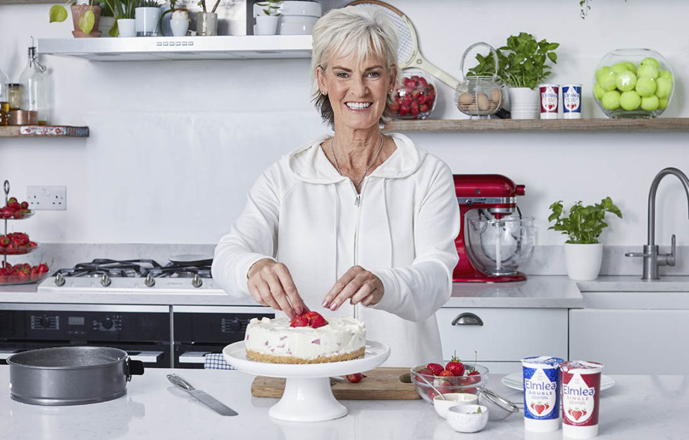 Judy Murray in kitchen, smiling, with strawberry cheesecake on worktop and plants, strawberries and jar of tennis balls on shelves behind