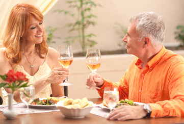 Mature couple at table in a brightly lit restaurant, raising glasses of white wine for a toast. She is wearing a strappy cream dress and he has a bright orange shirt.