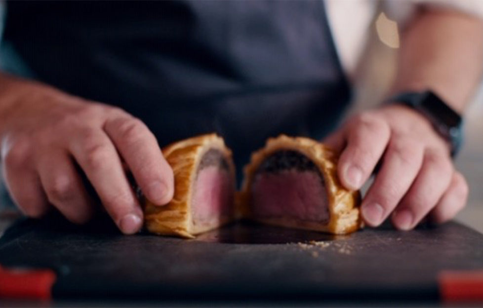 Chef has cur beef wellington in half to show pink meat, dark layer of mushrooms, and golden pastry