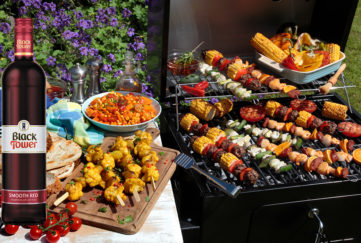 A barbecue grill filled with kebabs with bread and salad at the side and a bottle of Black Tower Smooth Red wine