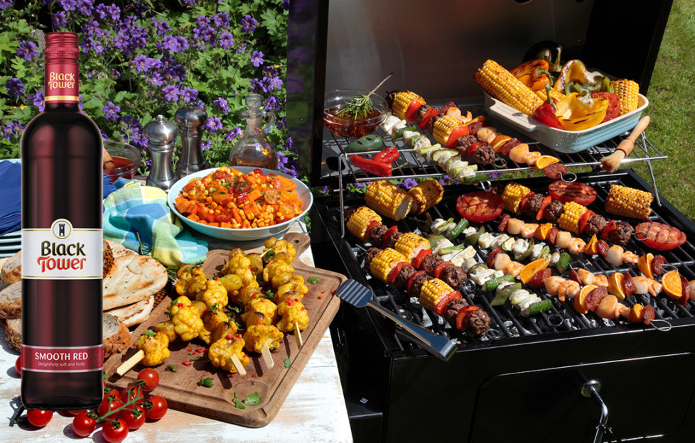 A barbecue grill filled with kebabs with bread and salad at the side and a bottle of Black Tower Smooth Red wine