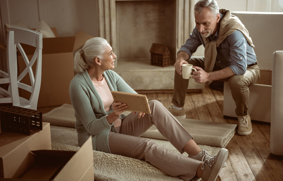 Older wife showing old photo to her husband while taking break from packing cardboard boxes