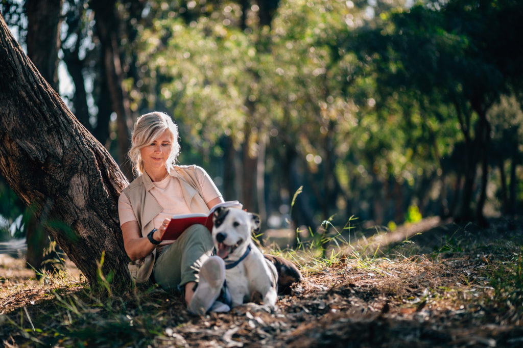Happy mature woman with dog sitting under forest tree and reading book in the woods