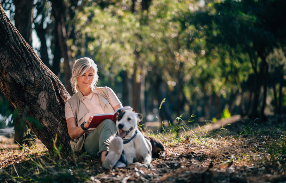 Happy mature woman with dog sitting under forest tree and reading book in the woods