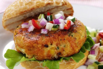 Open crusty white roll with golden brown salmon burger topped with diced red onion, red pepper and cucumbered with